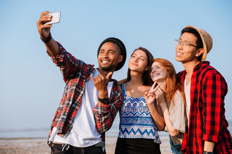Vier Personen machen ein Selfie von sich am Strand in der Sonne. Handy Akku sparen – Tipps für eine lange Akkulaufzeit unterwegs