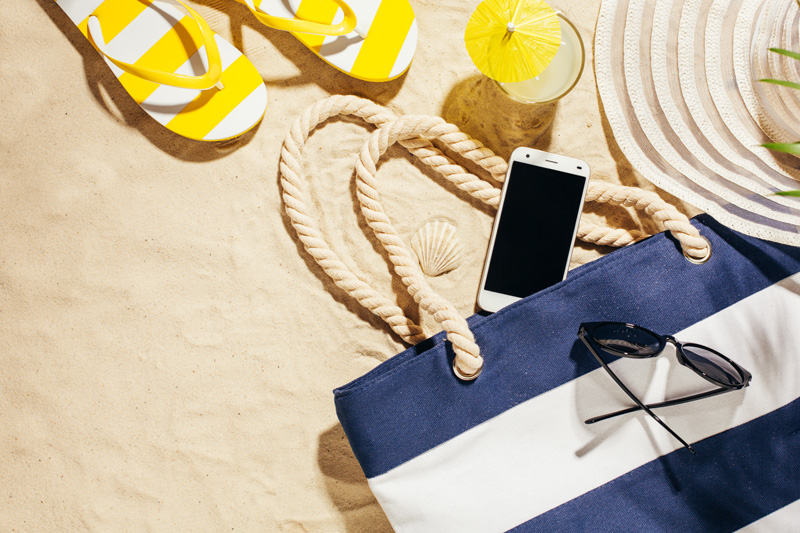 Handy liegt im Sand neben Strandtasche und Schuhen in der Sonne Handy Akku sparen: Schütze Dein Handy im Sommer vor der Sommerhitze!