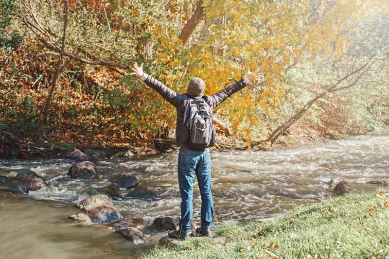 Ein Mann steht mit geöffneten Armen an einem Flussrand im Wald. Digital Detox schont nicht nur Deinen Handy Akku, sondern auch Deine Gesundheit!