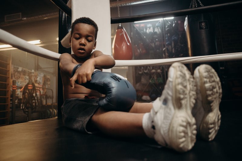 Ein im Boxring sitzender Junge zieht sich seine Boxhandschuhe an Im Boxring sitzender Junge zieht Boxhandschuhe für Training am Standboxsack an