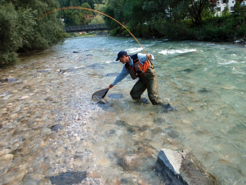 Ein Mann in Neopren Wathose mit Angel und Kescher angelt im Fluss.