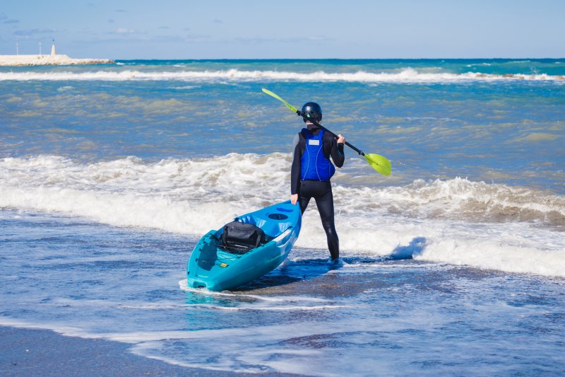 Paddelhose als Schutzbekleidung Kajakfahrer steht in Paddelhose mit Kajak und Paddel am Meer